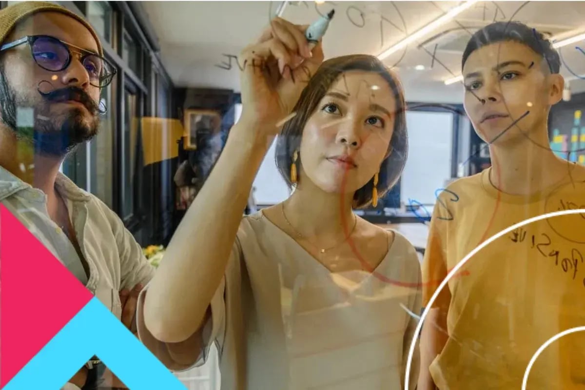 Three diverse coworkers collaborate in a bright office as a woman sketches on glass; teammates watch attentively, creating an innovative mood.