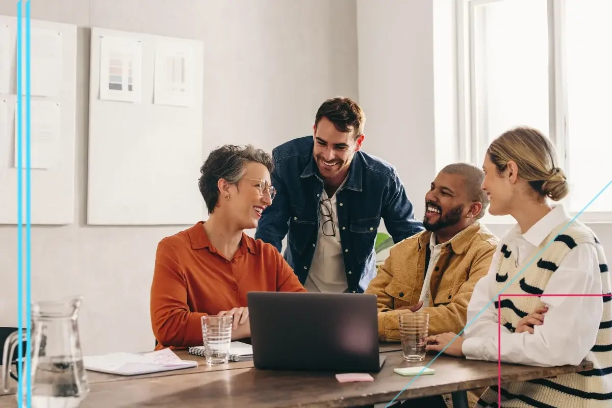 A diverse group of four smiling colleagues collaborate around a laptop at a wooden table in a bright office, fostering a positive and engaging atmosphere.