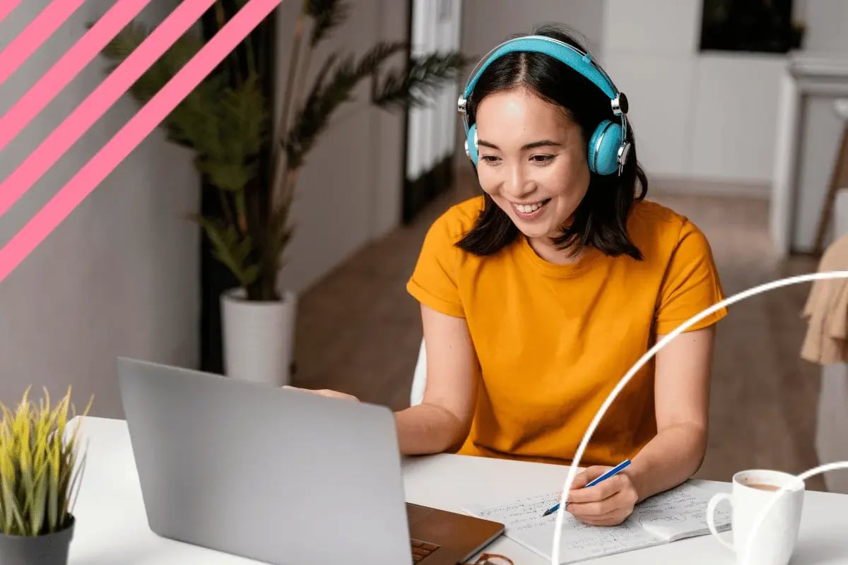 A woman in a yellow shirt with blue headphones smiles while using a laptop at a desk.
