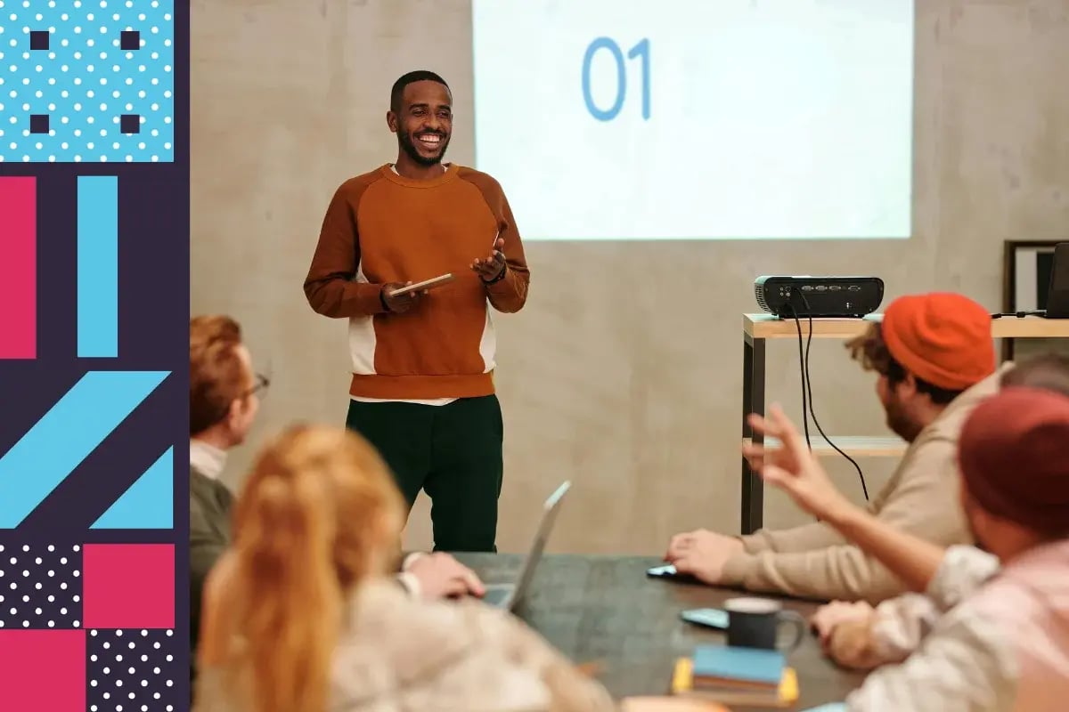 A man stands and smiles while presenting to a group seated at a table; a projected screen behind him displays the number 