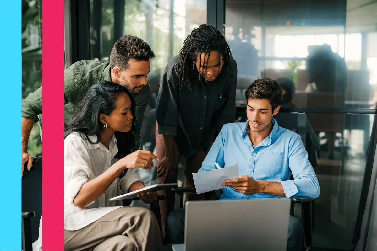A diverse group of four professionals collaborates around a laptop. One holds a document, guiding the discussion.