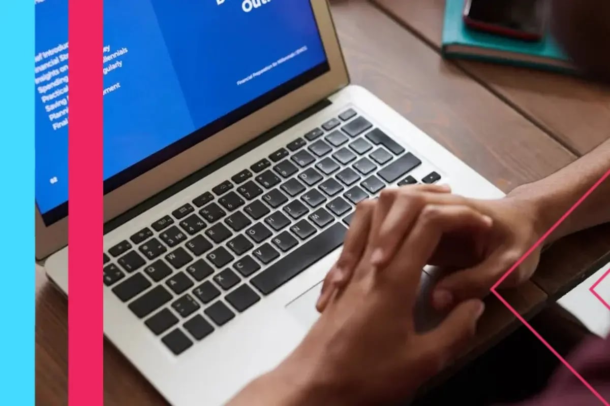 Close-up of hands typing on a laptop with blue coding text on screen, conveying focused work at a modern desk with pink graphic accents.