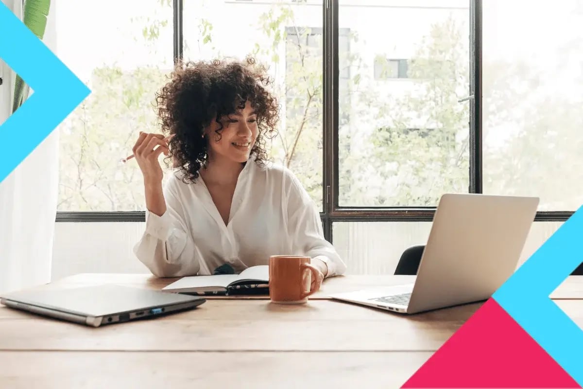 A woman with curly hair sits smiling at a laptop in a bright room. She holds a pen, with a notebook and mug on the table. The mood is relaxed and cheerful.