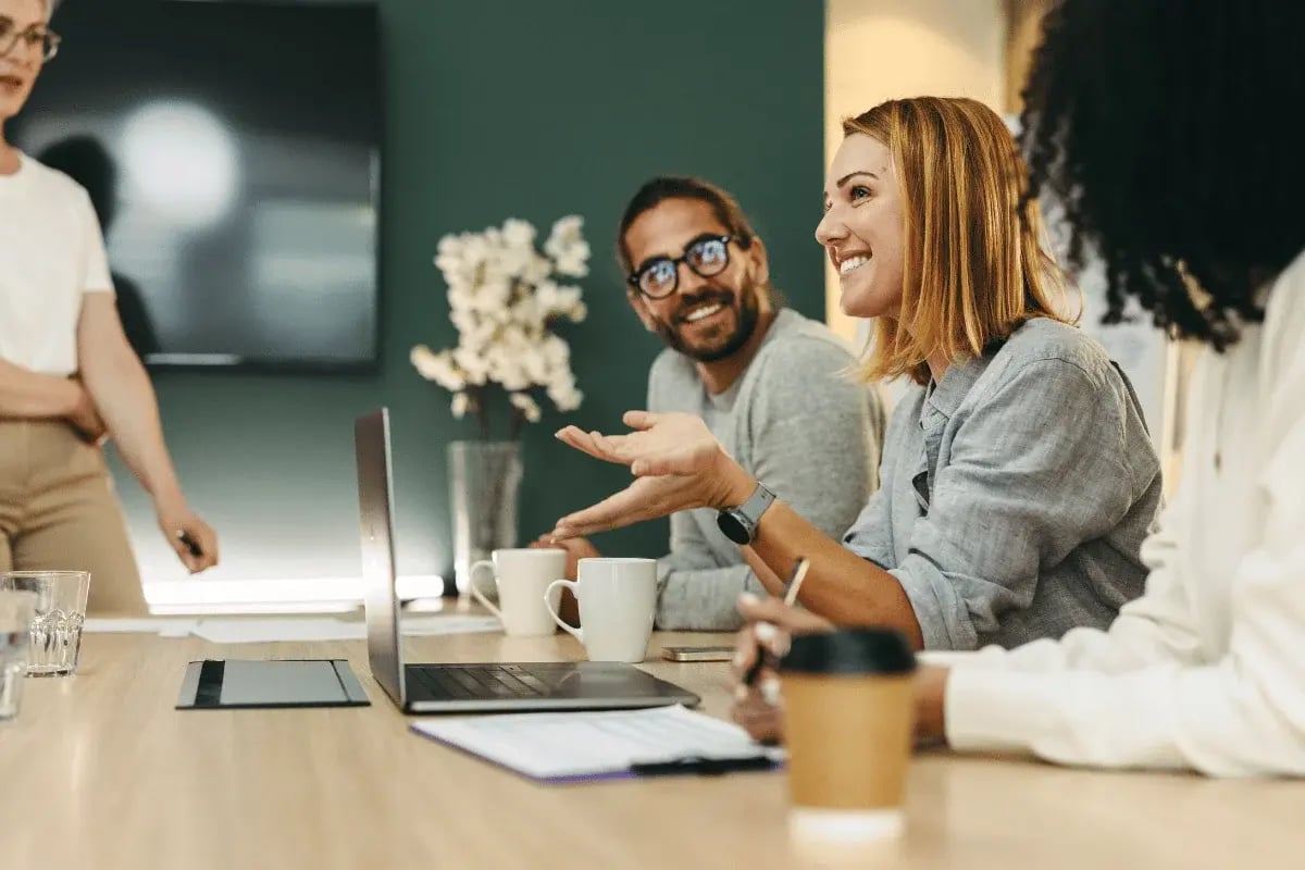 Three smiling colleagues sit at a table with laptops and coffee, engaged in discussion. One person gestures animatedly. Bright, collaborative atmosphere.