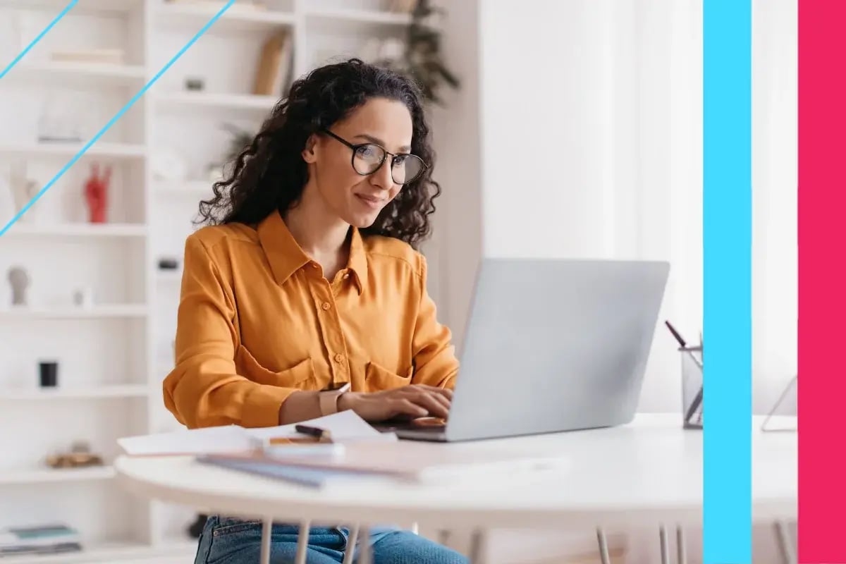 Smiling woman in glasses and an orange blouse works on a laptop at a bright home office desk, creating a focused, confident mood.