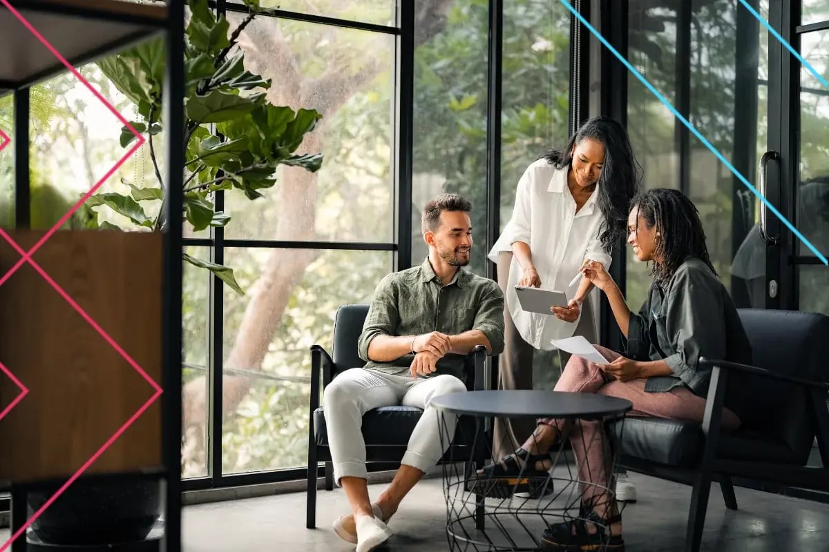 Three colleagues collaborate in a modern office with large windows, discussing documents and a tablet around a round black table.