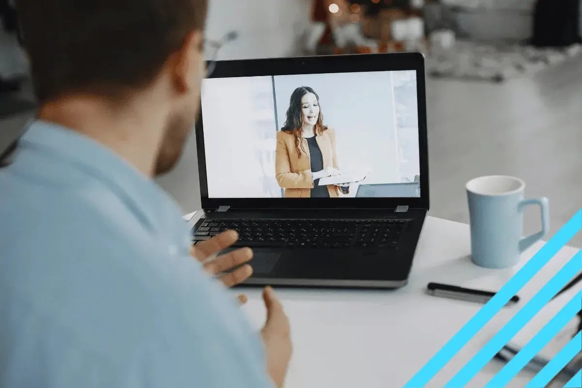 A man in a blue shirt participates in a video call on his laptop, showing a woman in a brown blazer presenting. A blue mug is on the desk.