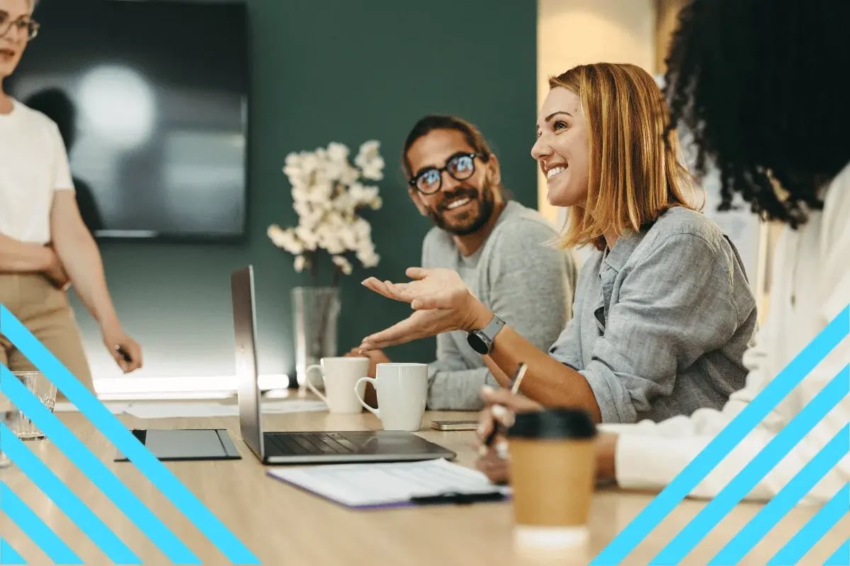 A woman cheerfully discusses at a table with laptops and coffee, while colleagues listen and smile. Collaborative atmosphere.