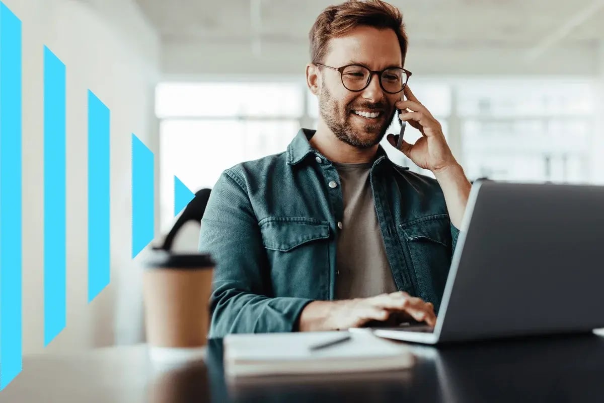 A man smiles while talking on a phone and using a laptop in a bright office.