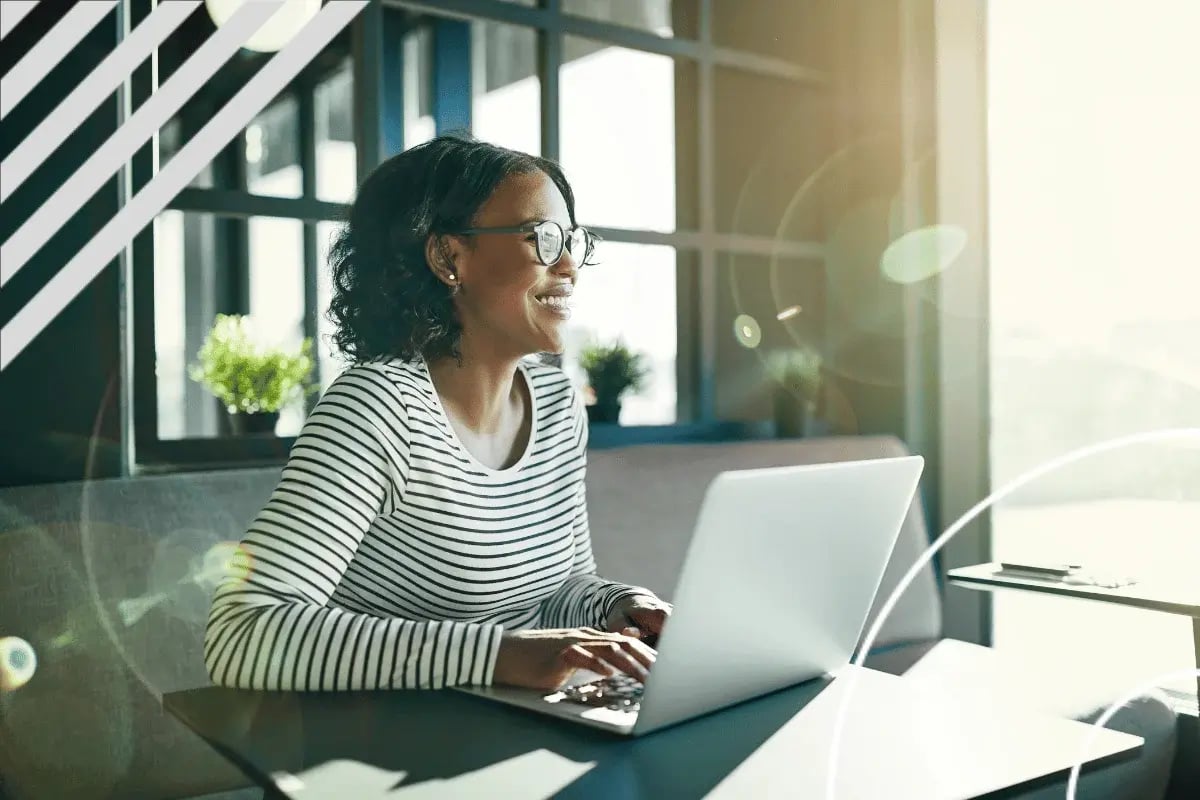 Person in a striped shirt typing on a laptop at a table with bright sunlight streaming through large windows behind them.