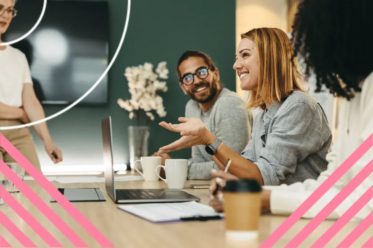 A woman smiles and gestures during a meeting, surrounded by colleagues around a table with laptops and coffee mugs. 