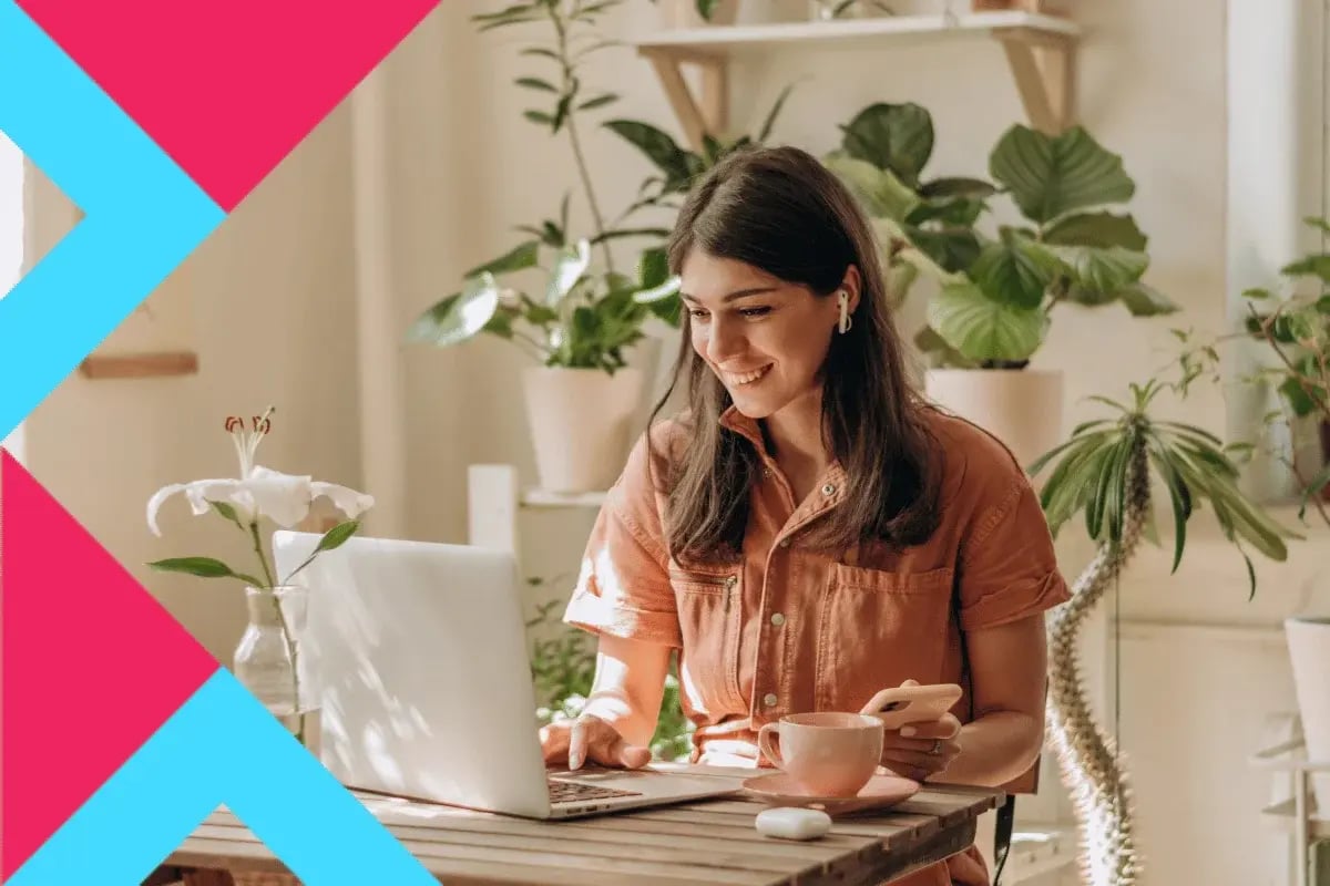 A woman in a peach shirt, wearing earbuds, smiles while working on a laptop at a wooden table.