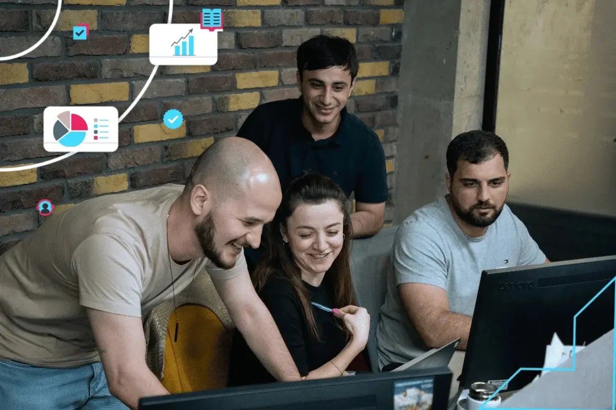 Four people gather and smile at a computer screen, surrounded by floating digital charts and icons, suggesting a collaborative, tech-focused environment.