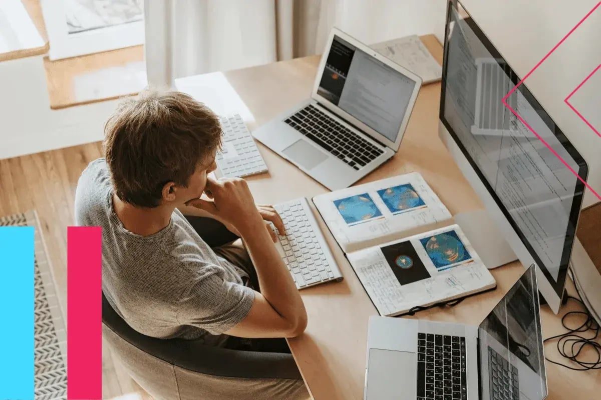 A person sits at a desk with multiple laptops and a large monitor, intensely focused. Open books and digital coding display a tech-focused workspace ambiance.