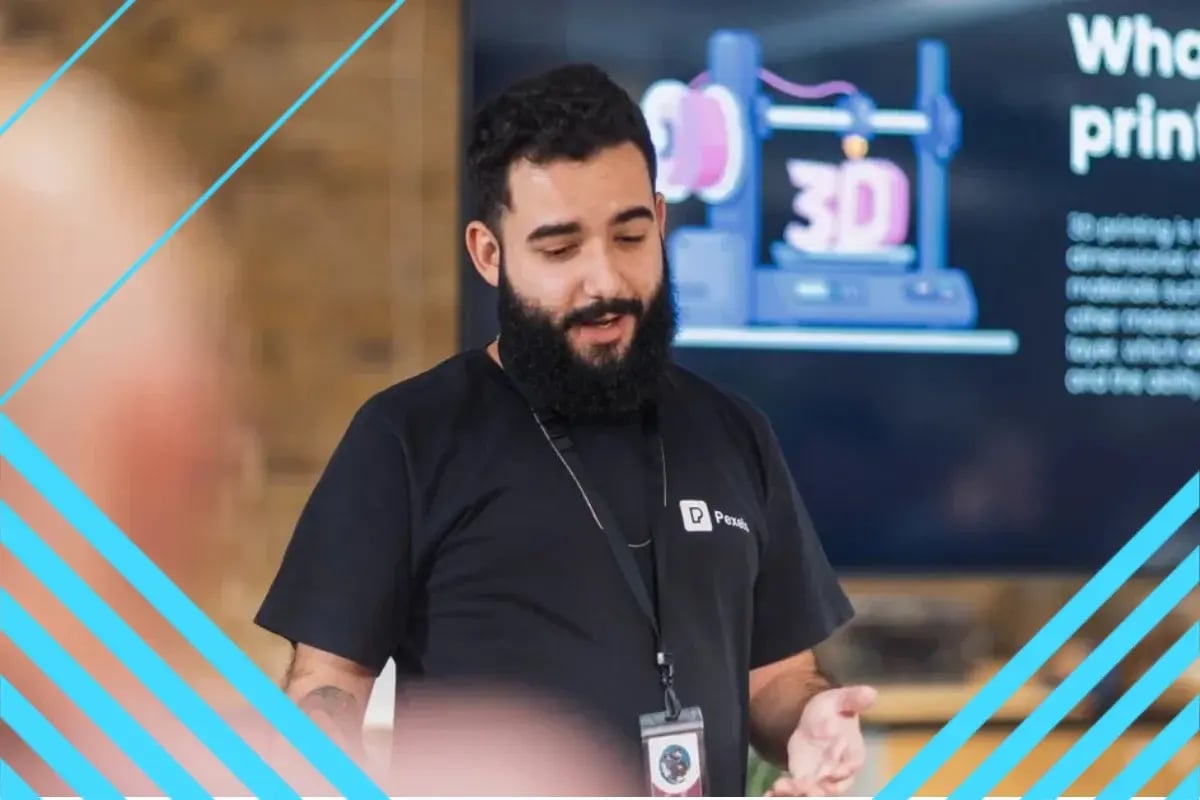 Man with beard and black shirt giving a presentation about 3D printing with screen and audience in background.