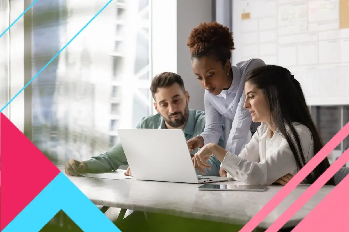 Three coworkers collaborate over a laptop in a bright office, discussing work with focused, and engaged expressions.