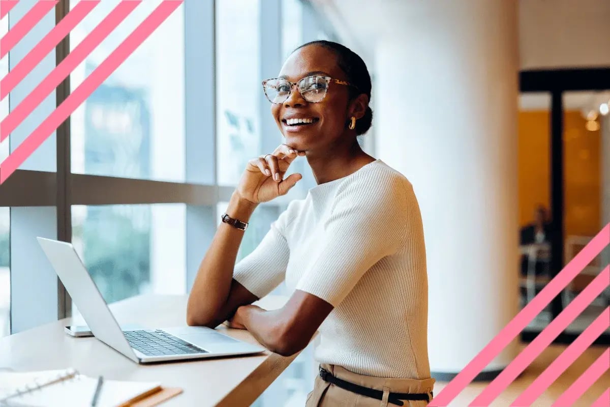 Smiling woman in glasses sits at a desk with a laptop, looking out a window.