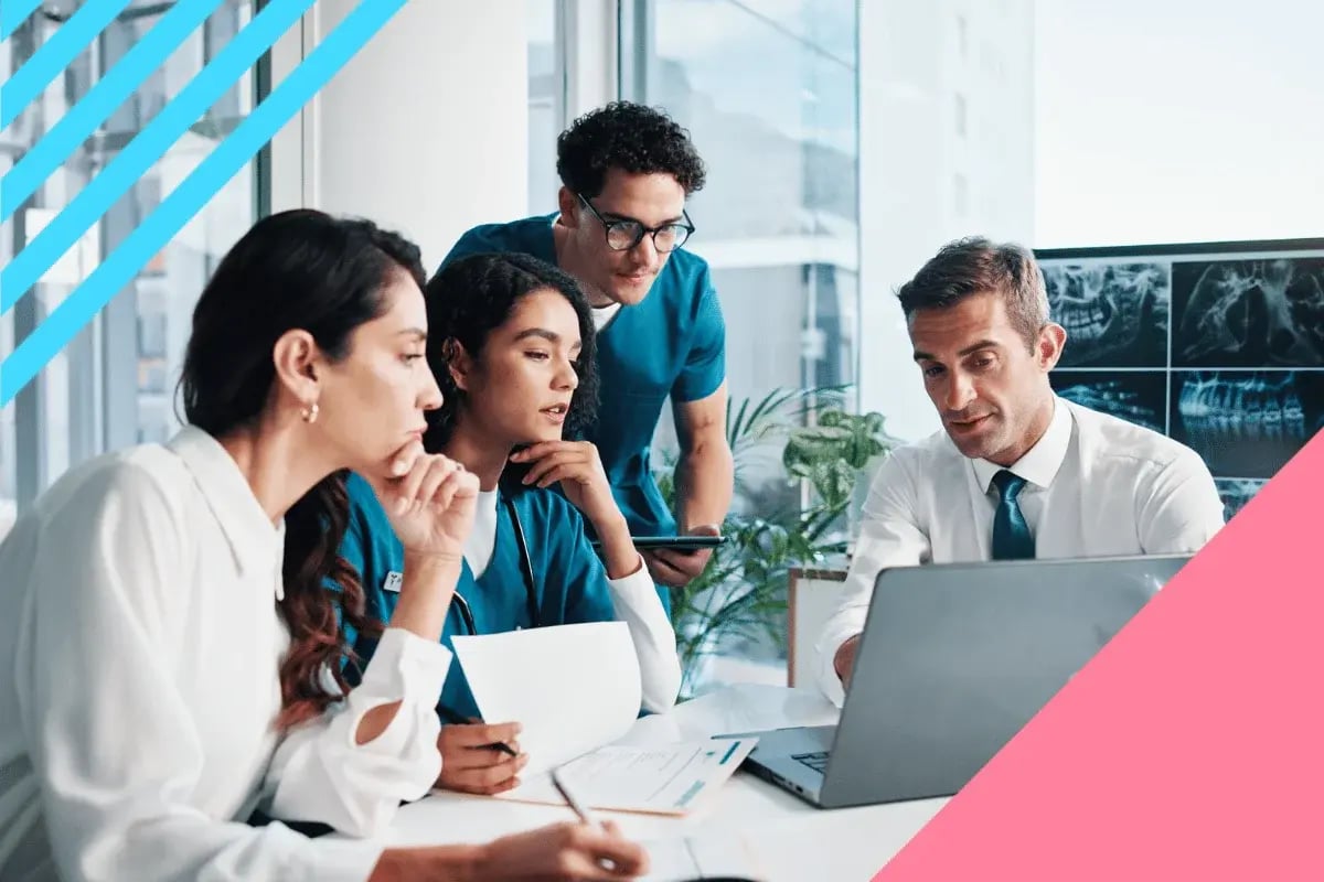 A diverse group of healthcare professionals in a meeting, focused on a laptop. They appear engaged and collaborative.