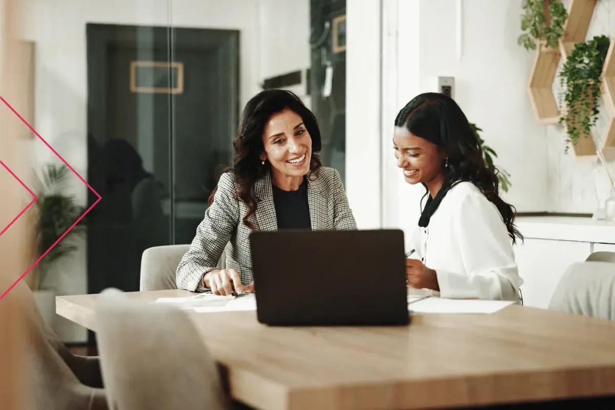 Two women engaged in a friendly discussion at a table with a laptop, surrounded by modern decor and plants, conveying a professional, collaborative vibe.
