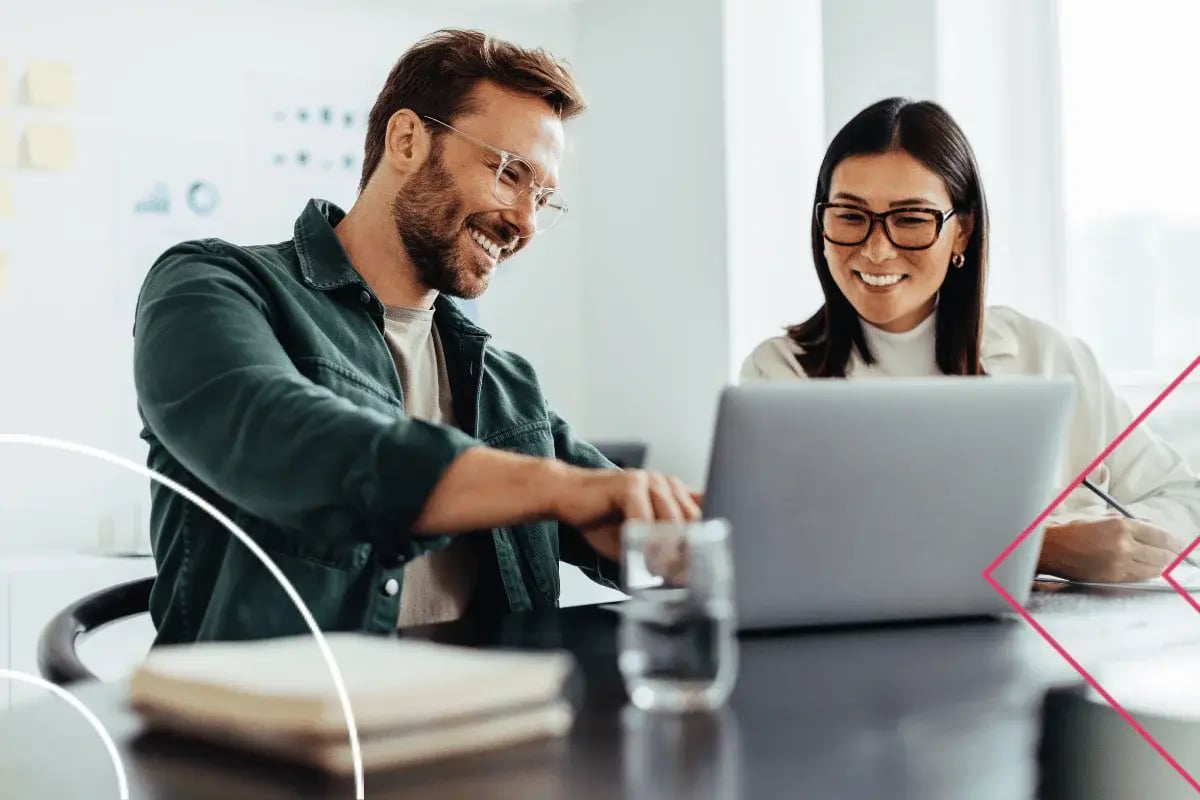 A man and woman smile while working together on a laptop in a bright office. They appear focused and collaborative, conveying a positive, professional atmosphere.