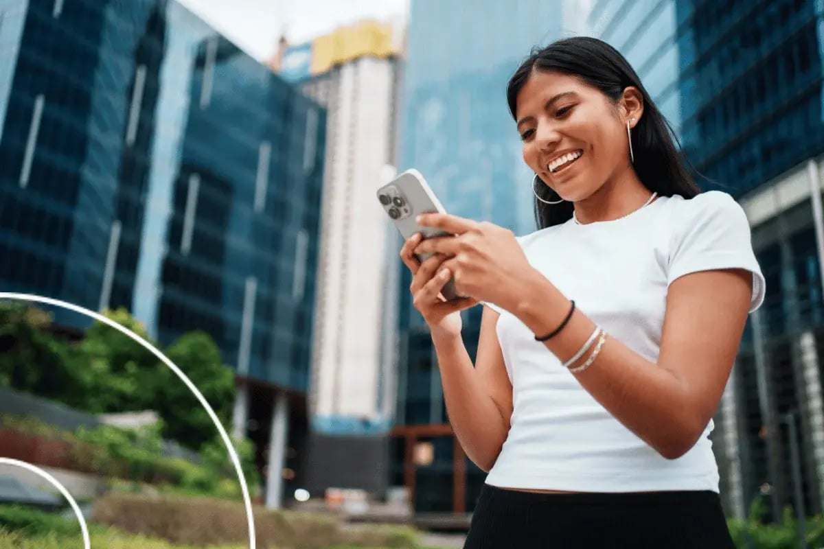 A person in a white shirt stands outdoors, looking at their smartphone, with modern buildings in the background.