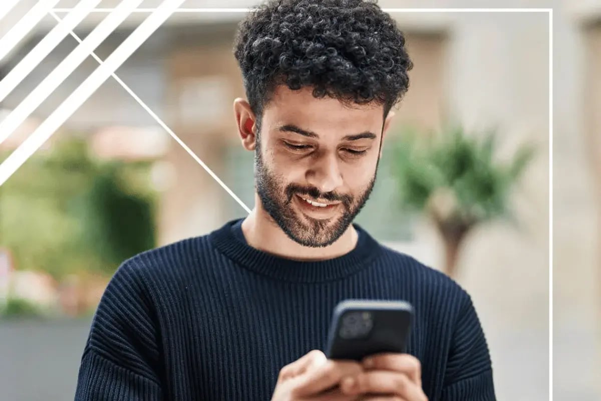 A person with curly hair is holding a smartphone, standing outdoors with greenery in the background.