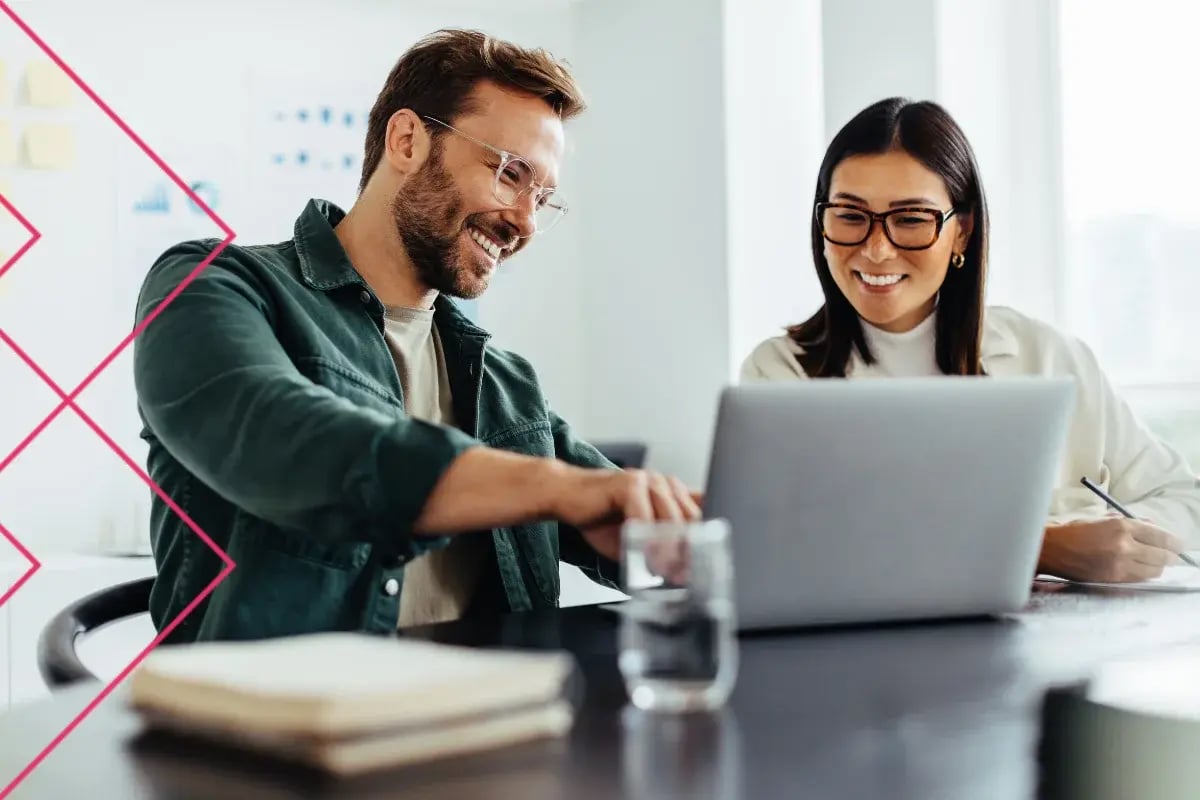 Smiling coworkers collaborate at a laptop in a bright office, with a man gesturing and a woman taking notes; friendly, productive mood.