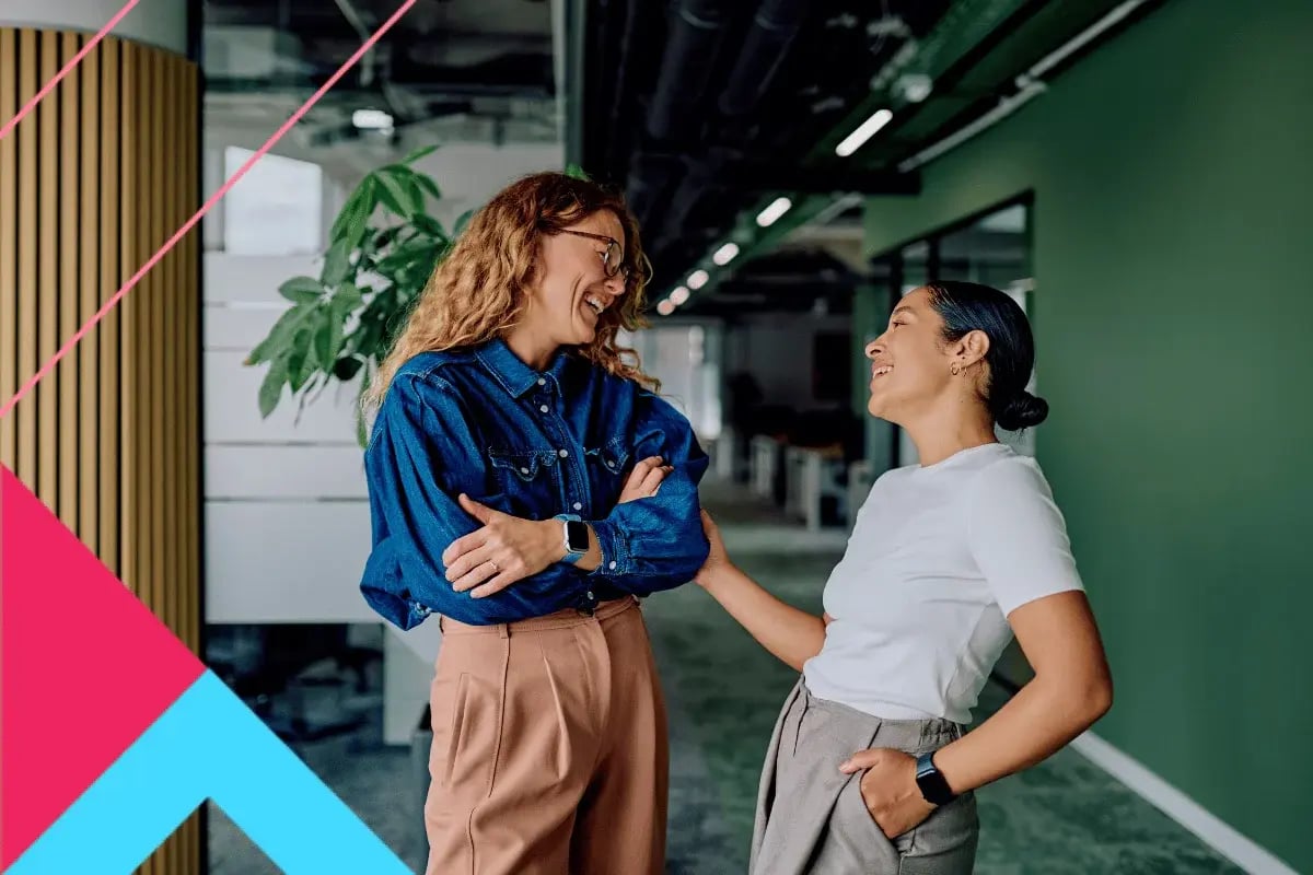 Two women in casual clothing share a laugh in a modern office with green walls and plants. The atmosphere is friendly and relaxed.