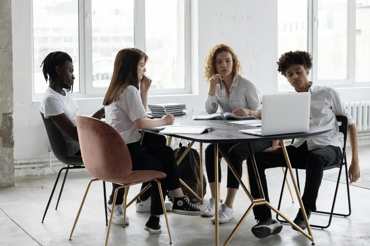 A group of five people engaged in a discussion around a modern table with books and a laptop in a bright, airy room.