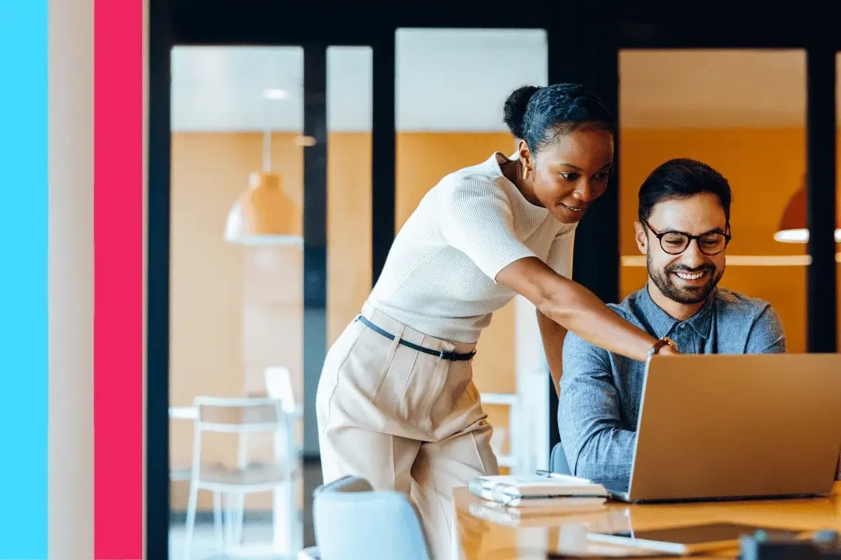 A woman and a man are in an office. The woman points at a laptop screen while the man smiles, suggesting teamwork and collaboration.