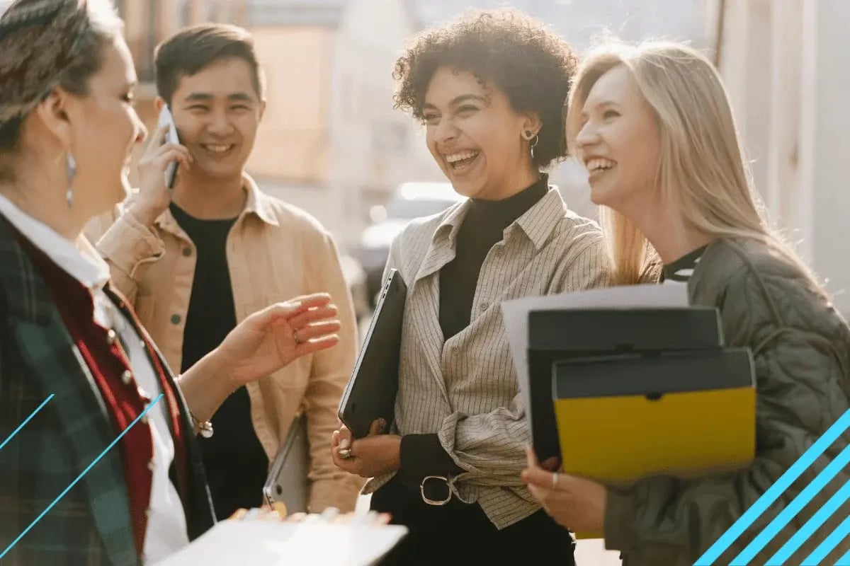Four diverse young adults smiling and chatting outdoors in sunlight. One person is on the phone, while others hold folders, creating a joyful atmosphere.