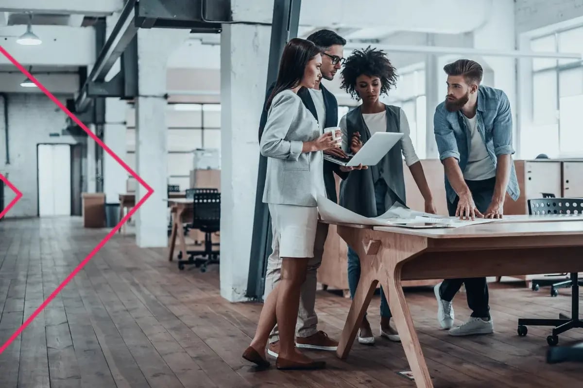 A diverse group of four professionals collaborates in a modern office, gathered around a wooden desk with a laptop and papers, conveying teamwork and focus.