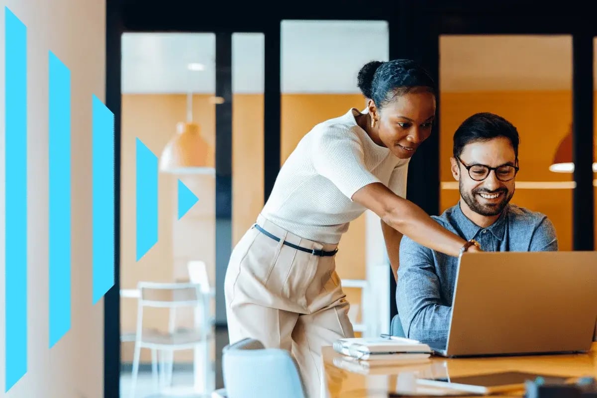 A woman points at a laptop screen while helping a smiling man in an office setting. Blue arrow graphics are on the left, suggesting progress.