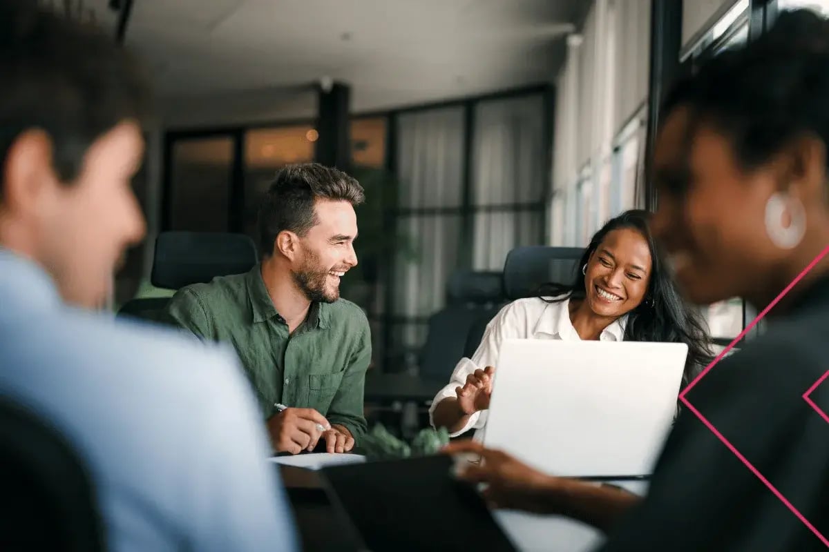 A group of diverse professionals in a modern office, gathered around laptops, smiling and engaged in conversation. The atmosphere is collaborative and positive.