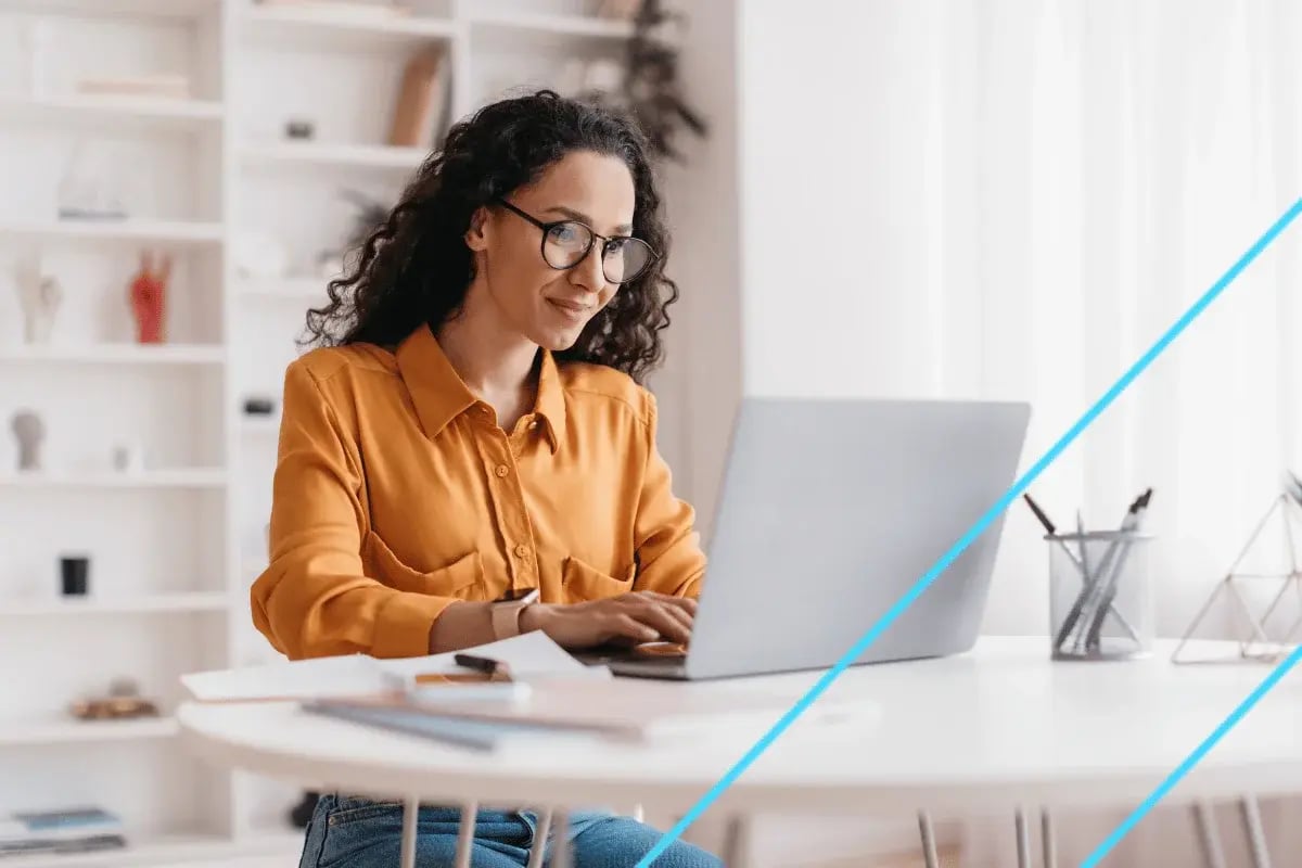 A person types on a laptop at a bright, modern workspace, featuring a white table and decorative shelves in the background.