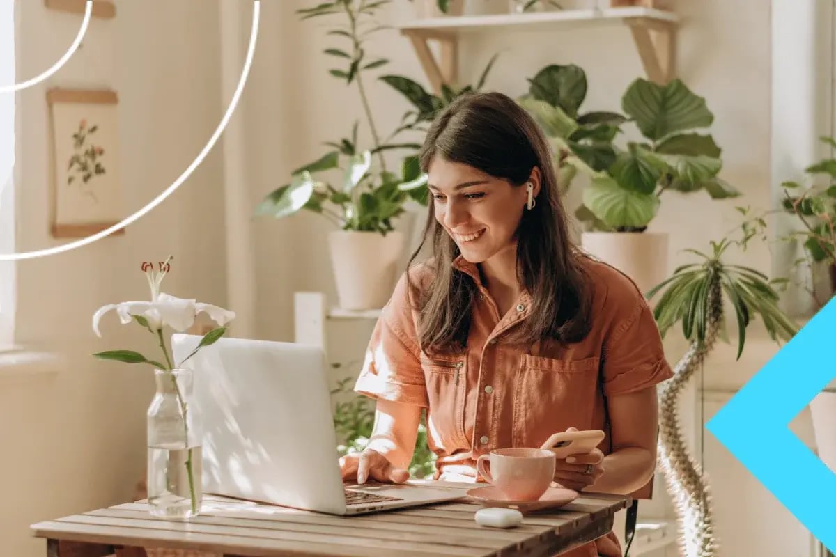 A person sits at a wooden table with a laptop, phone, and a pink cup, surrounded by green plants and soft natural light.