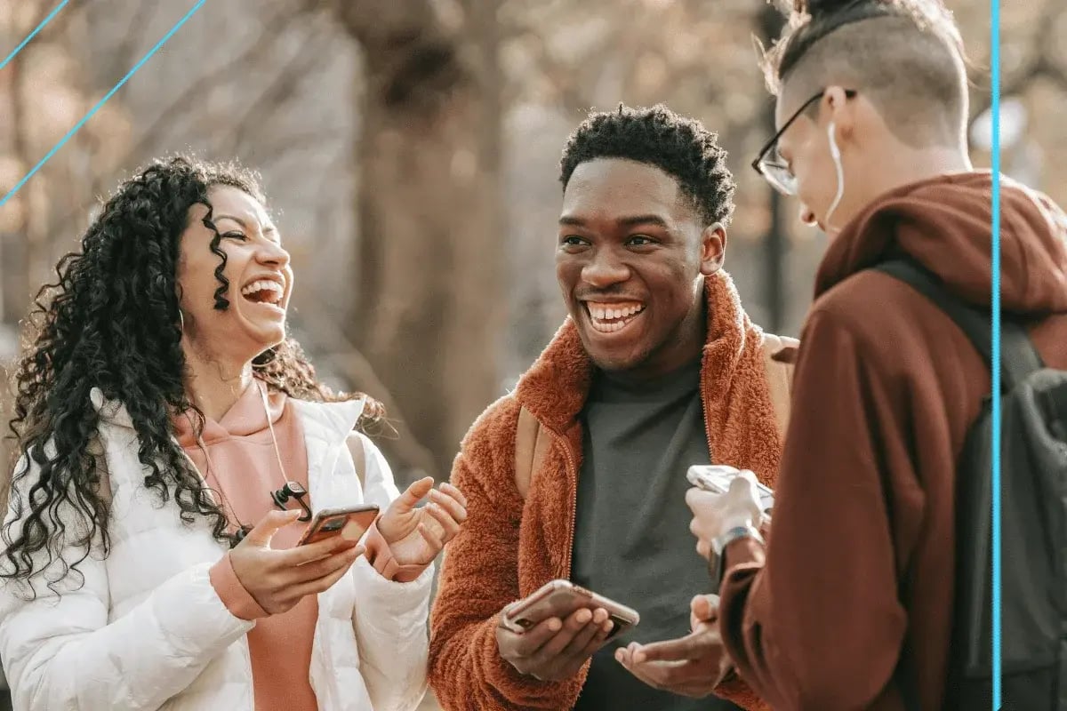 Three friends outdoors laughing and chatting, holding smartphones. 