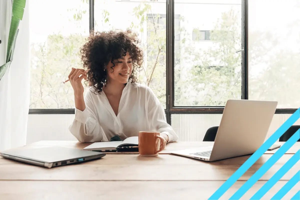 Woman working on laptop at bright desk with notebook, coffee mug, and large window.