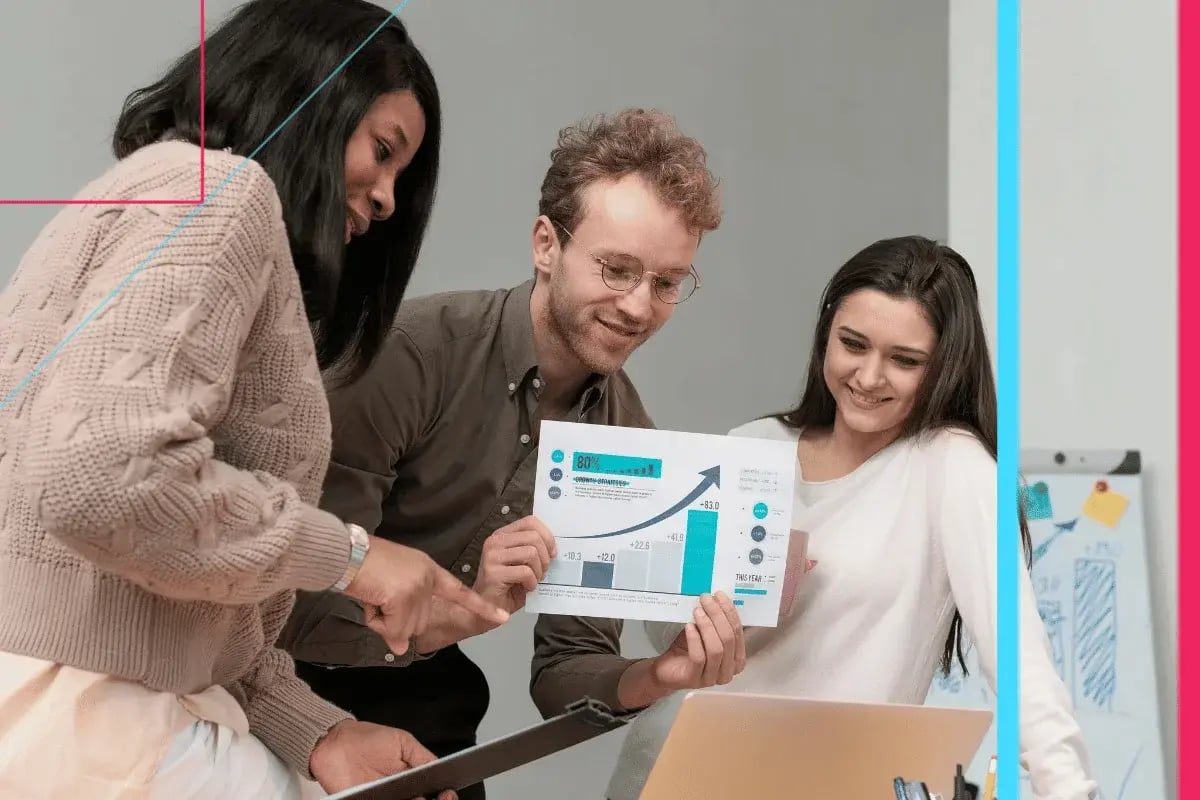 Three colleagues smiling, engaged in a discussion around a laptop, as one holds a graph, suggesting a collaborative and positive work environment.