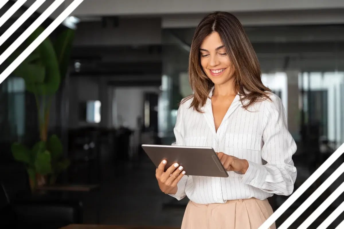 A smiling woman in a white blouse uses a tablet in a modern office.