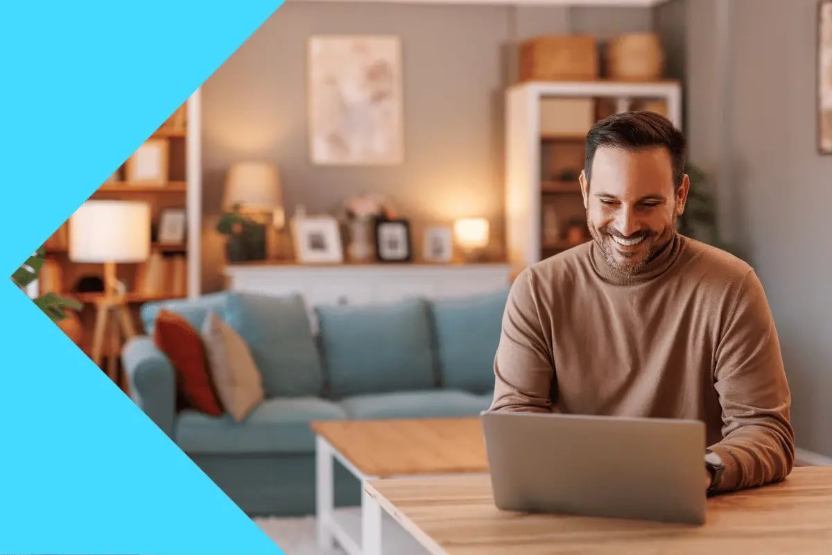 A man in a cozy living room setting is smiling at his laptop. 