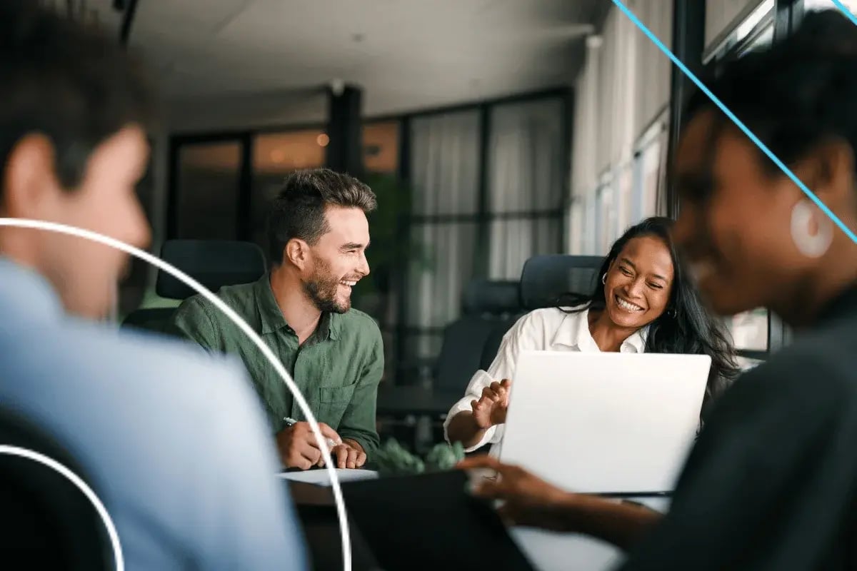 Team members smiling during a collaborative office meeting around a laptop, discussing ideas in a modern conference room.