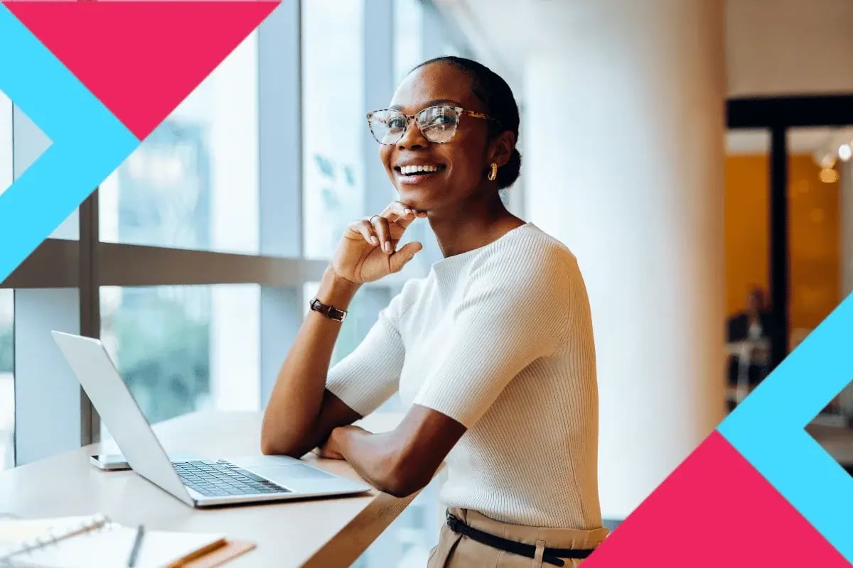 Smiling woman with glasses using a laptop at a modern desk, thoughtfully looking out a bright office window.