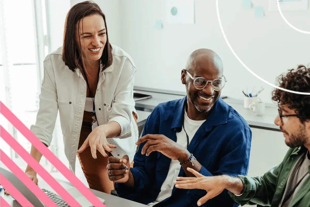 A diverse group of three colleagues, smiling and engaged in discussion at a desk, conveying a collaborative and positive work environment.