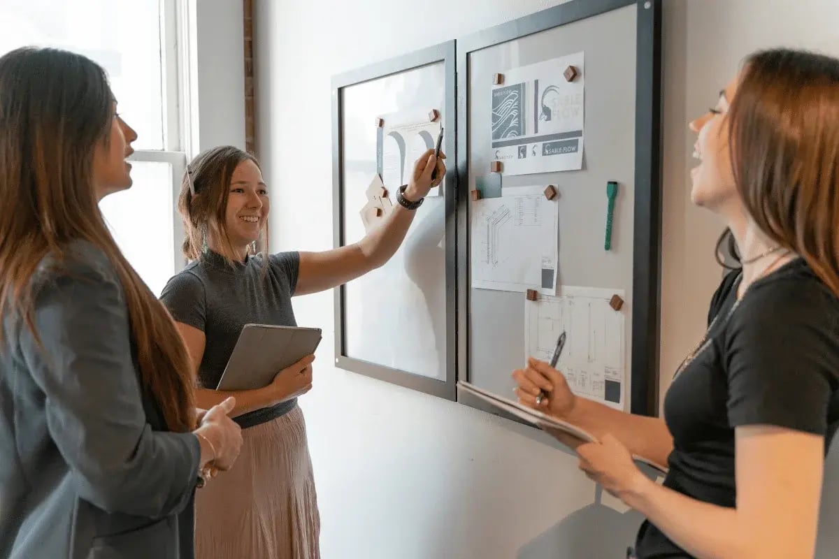 Three women stand by a wall-mounted whiteboard covered with papers, discussing and writing notes during a meeting.