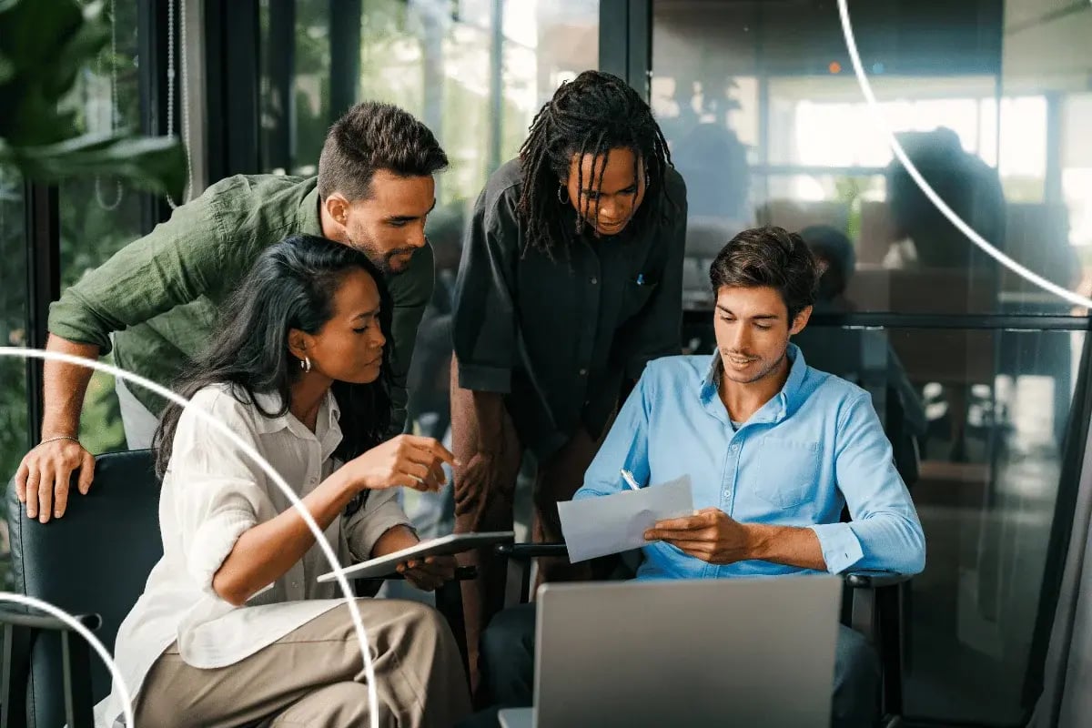 A diverse group of four colleagues engaged in a collaborative meeting, gathered around a laptop. They are focused and discussing a document.