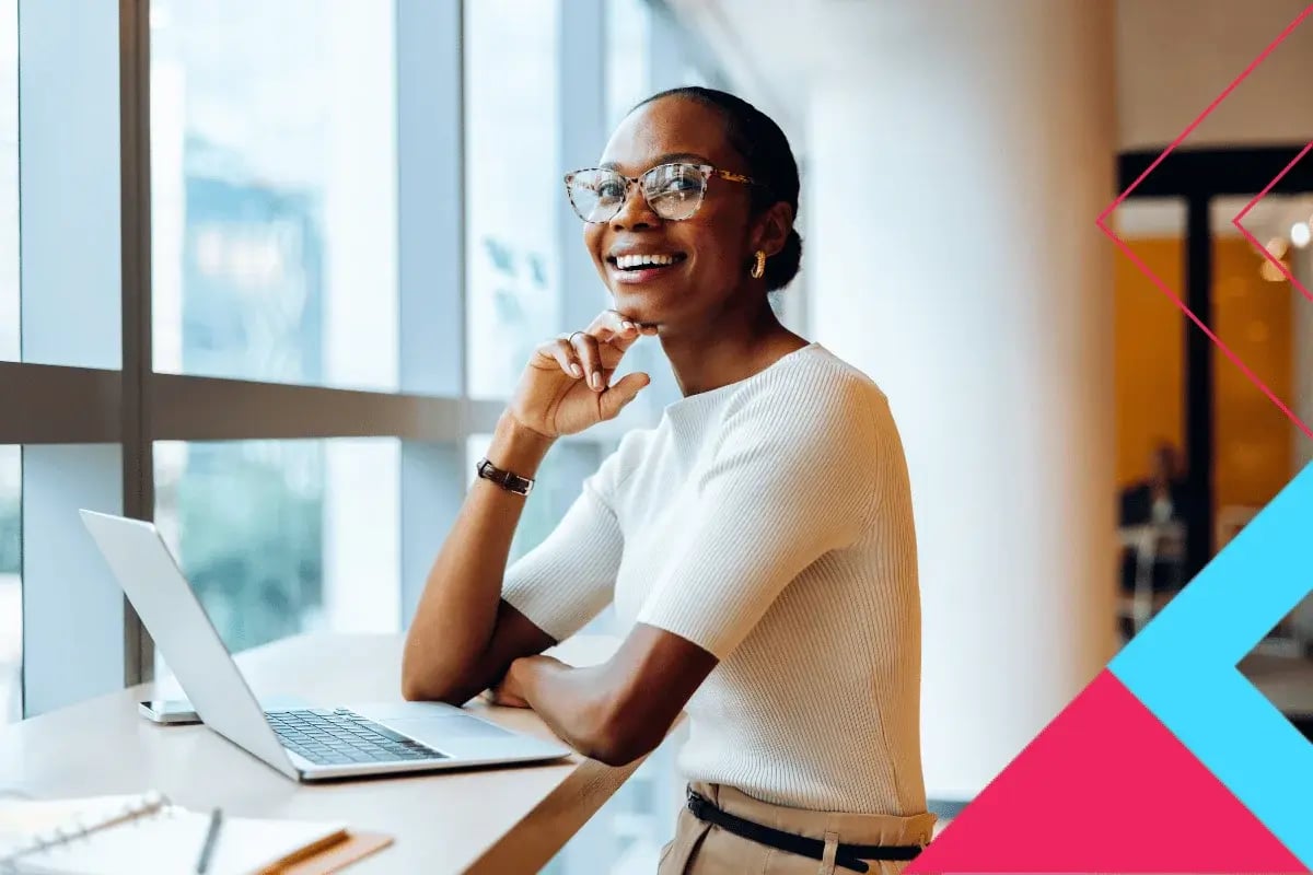 A person in a white top sits thoughtfully at a desk with a laptop, looking out a large window with a modern interior backdrop.