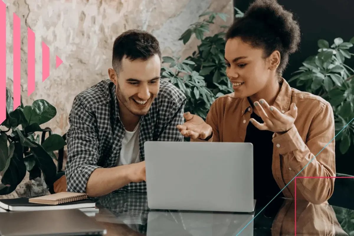 Two people sit at a table, smiling and gesturing at a laptop screen.