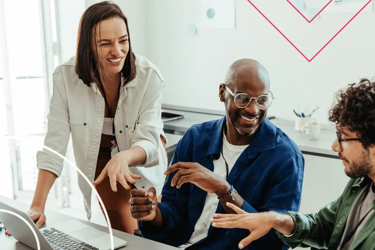 Three colleagues, two men and one woman, happily collaborate at a table with a laptop in a bright office.