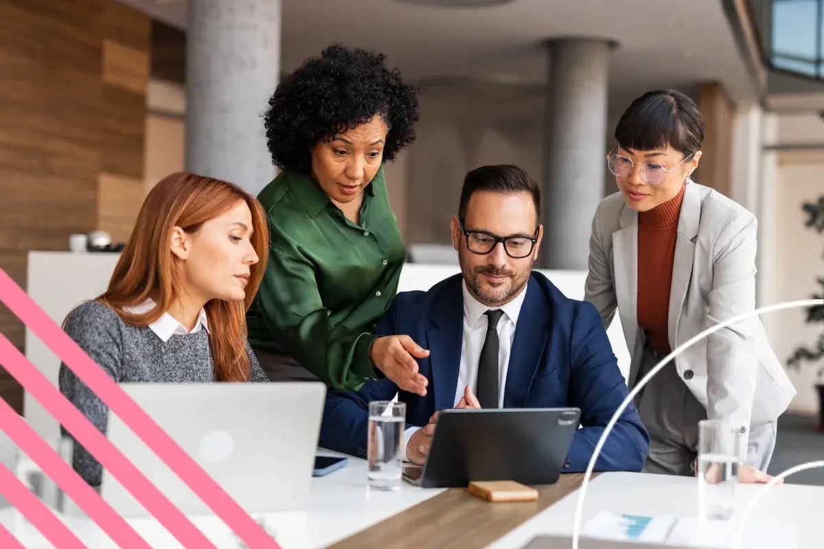A diverse group of four professionals engaged in a focused discussion around a tablet in a modern office setting, conveying teamwork and collaboration.